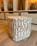 A square upholstered ottoman with a cream fabric and a brown geometric line pattern, placed on a light wood floor in front of a beige cabinet.